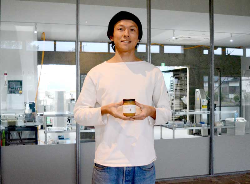 新工場を背景にピーナツバターを手に笑みを浮かべる中野代表＝山武市