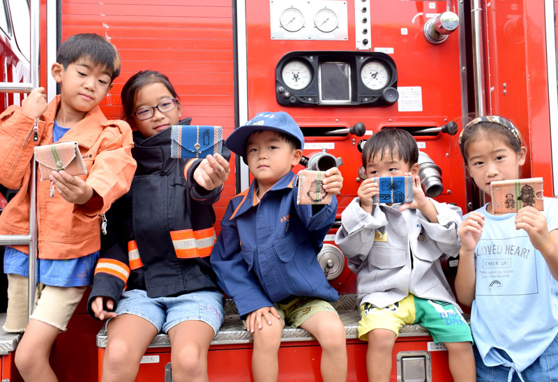 防火衣や救助服を着て完成した小銭入れを見せる子どもたち