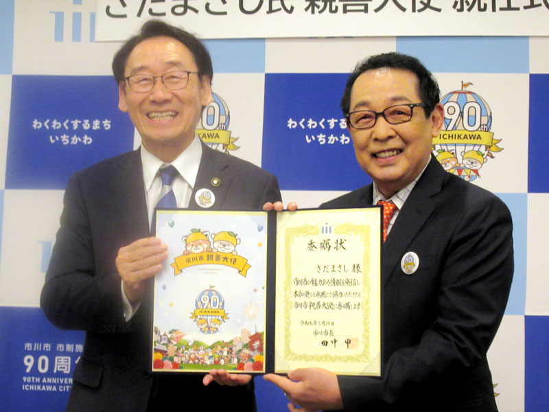 親善大使の委嘱状を見せるさださん。左は田中甲市長＝５月、市川市文化会館